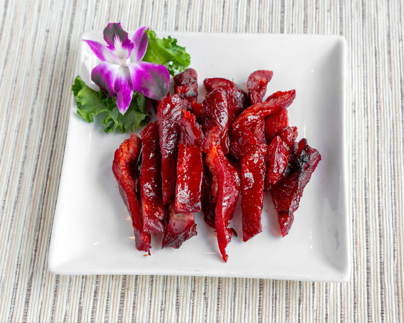 Bright red char siu pork slices on a white rectangular plate, garnished with a purple orchid and lettuce.