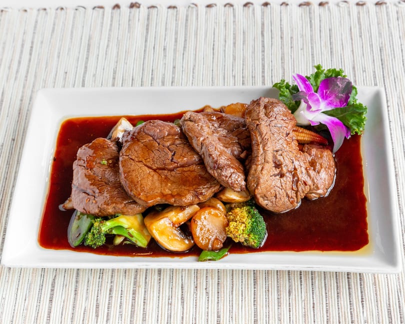 Beef medallions with broccoli, mushrooms, baby corn, and a purple orchid garnish in a rich brown sauce.