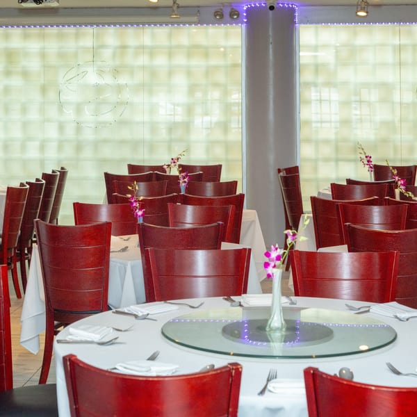 An empty dining room with many round tables set with white tablecloths and red wooden chairs.