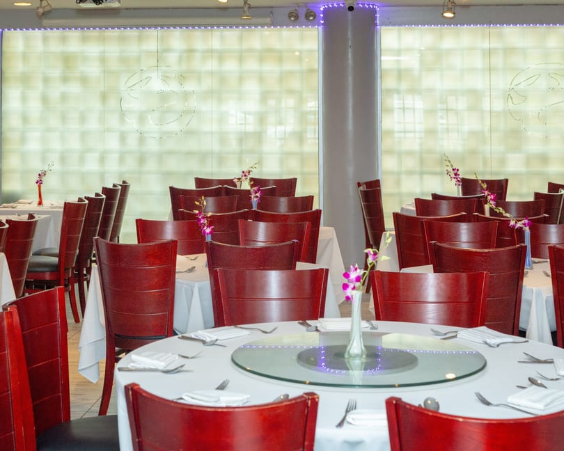 An empty dining room with many round tables set with white tablecloths and red wooden chairs.