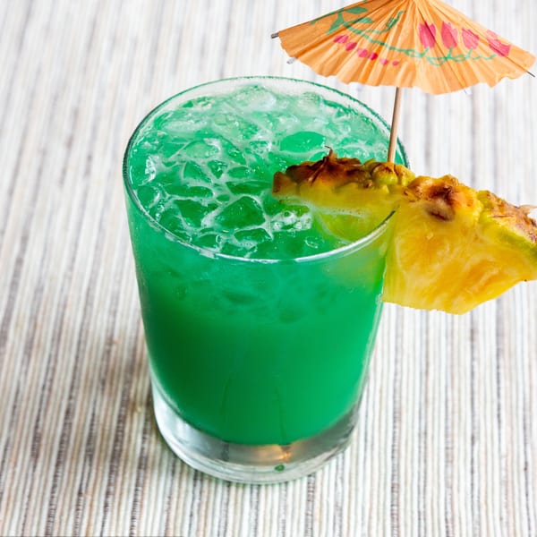 Vibrant green cocktail with ice, garnished with a pineapple wedge and a small paper umbrella.