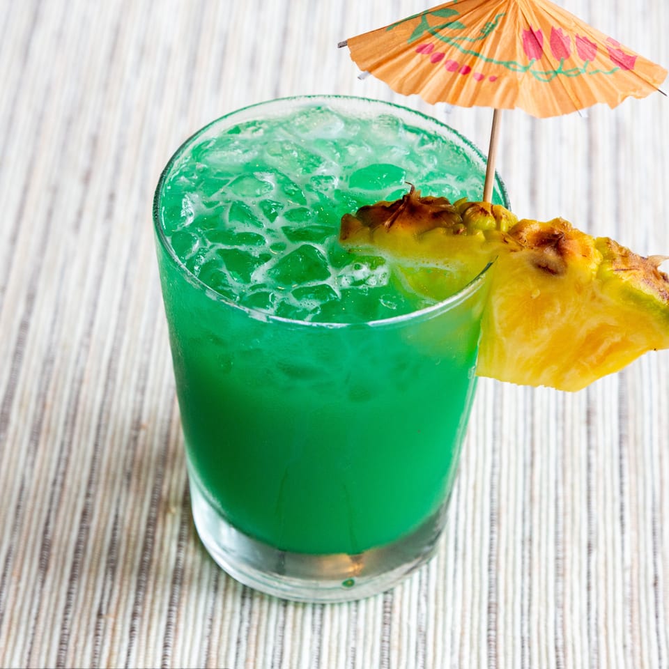 Vibrant green cocktail with ice, garnished with a pineapple wedge and a small paper umbrella.