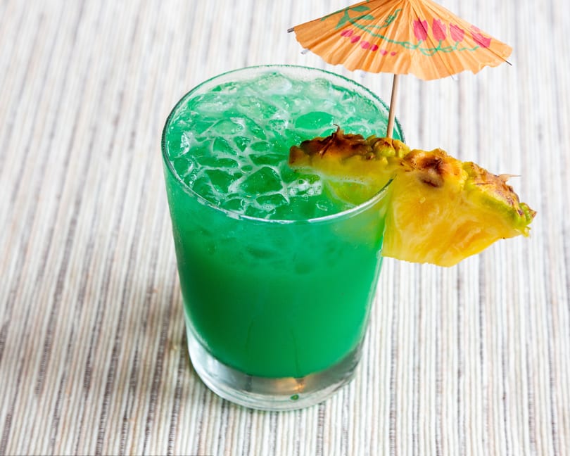 Vibrant green cocktail with ice, garnished with a pineapple wedge and a small paper umbrella.