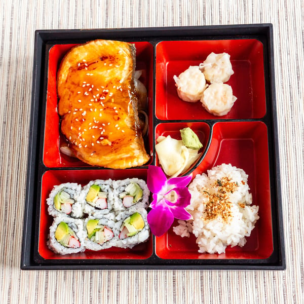 A bento box with teriyaki salmon, shrimp shumai, California rolls, rice, and a purple orchid.