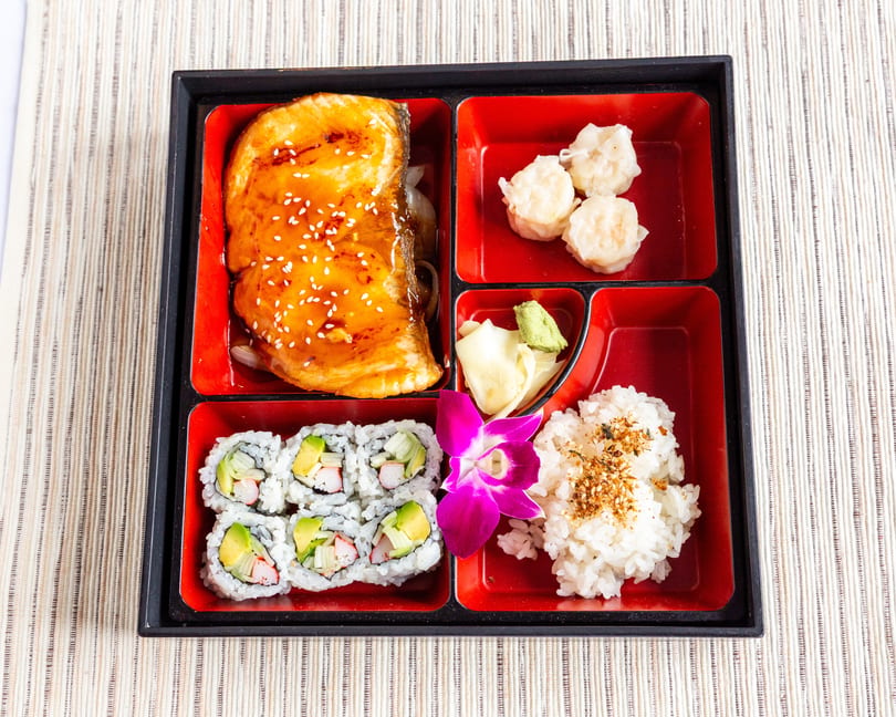 A bento box with teriyaki salmon, shrimp shumai, California rolls, rice, and a purple orchid.