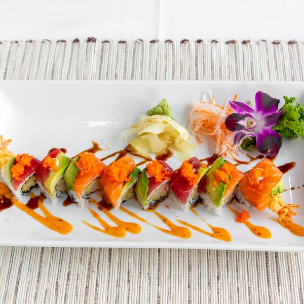A long white plate of colorful sushi rolls with sauces, wasabi, ginger, and an orchid garnish.