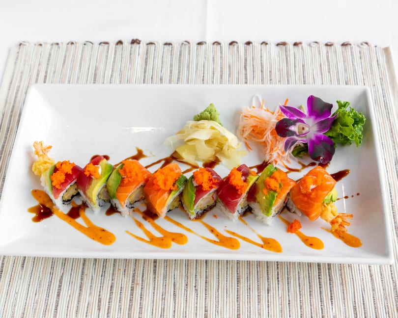 A long white plate of colorful sushi rolls with sauces, wasabi, ginger, and an orchid garnish.