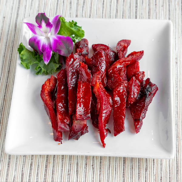 Bright red char siu pork slices garnished with a purple orchid and lettuce on a white plate.
