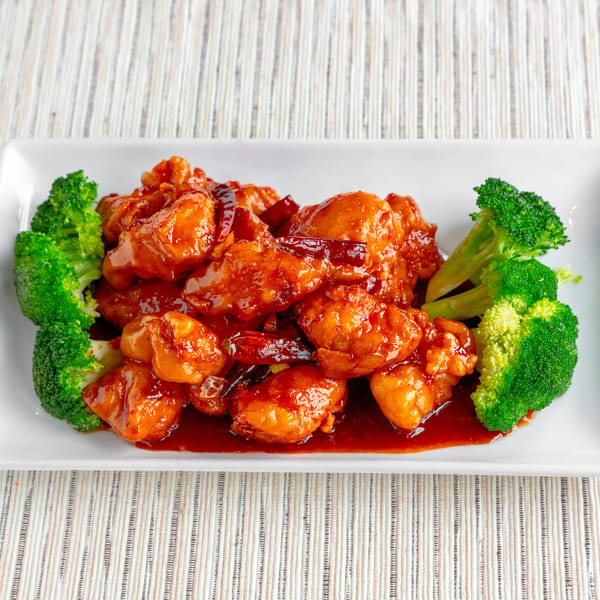 General Tso's chicken with dried red chilies and broccoli florets on a white rectangular plate.