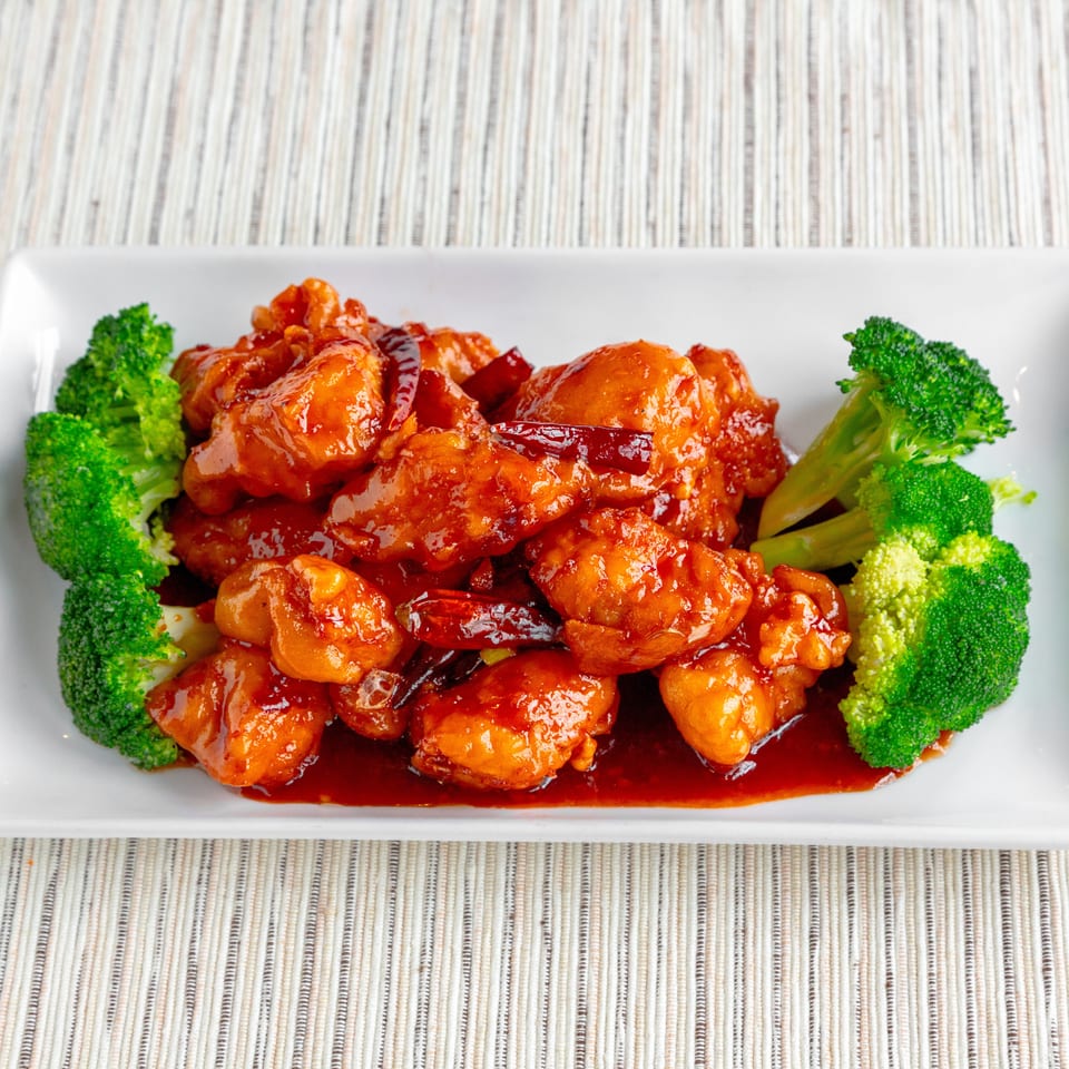 General Tso's chicken with dried red chilies and broccoli florets on a white rectangular plate.