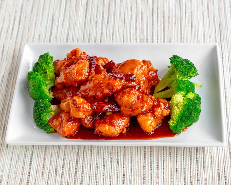 General Tso's chicken with dried red chilies and broccoli florets on a white rectangular plate.