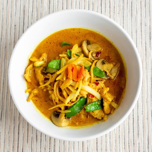 A white bowl filled with yellow curry, chicken, mushrooms, onions, and green peppers.