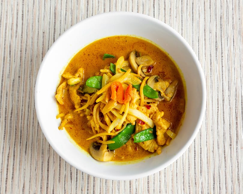 A white bowl filled with yellow curry, chicken, mushrooms, onions, and green peppers.