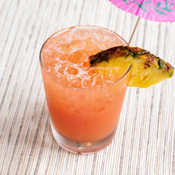 A vibrant orange cocktail with ice, garnished with a pineapple wedge and a pink umbrella.
