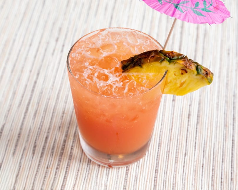 A vibrant orange cocktail with ice, garnished with a pineapple wedge and a pink umbrella.