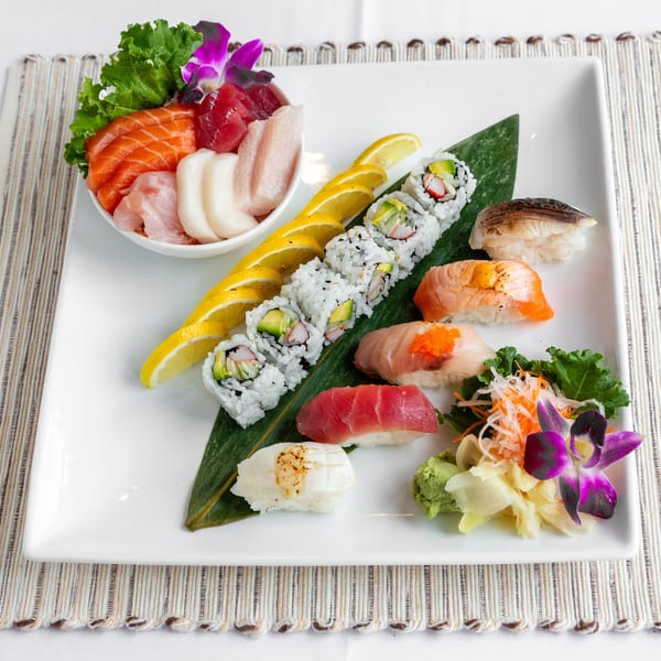 A white rectangular plate displays a variety of sushi, sashimi, and rolls, garnished with lemon slices and orchids.