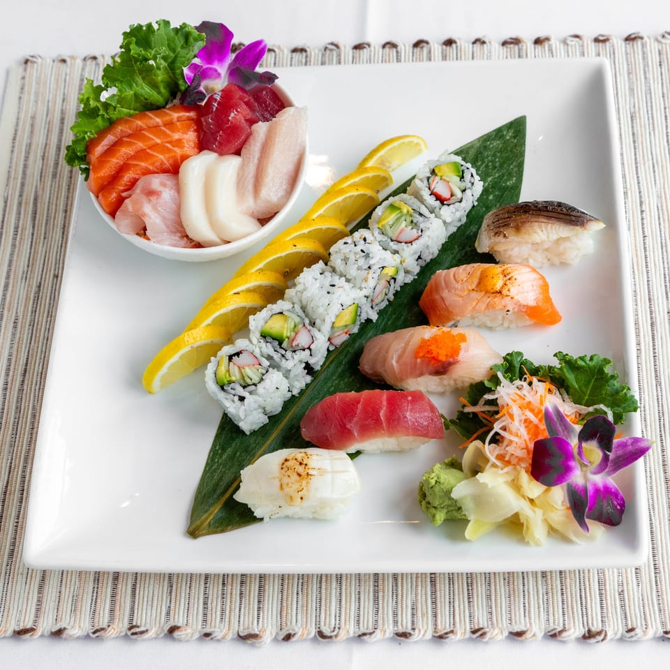 A white rectangular plate displays a variety of sushi, sashimi, and rolls, garnished with lemon slices and orchids.