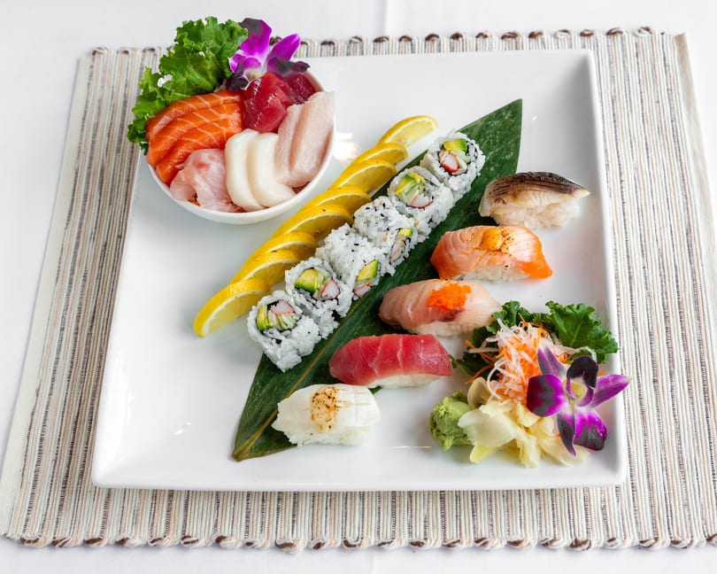A white rectangular plate displays a variety of sushi, sashimi, and rolls, garnished with lemon slices and orchids.