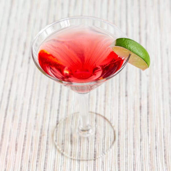 Vibrant red cocktail in a martini glass, garnished with a fresh lime wedge on a striped surface.