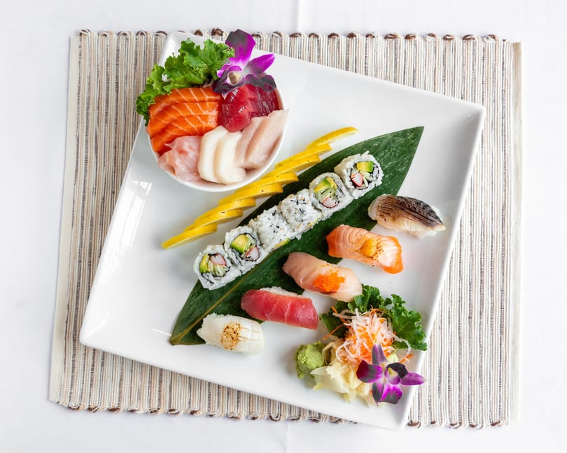 A white rectangular plate holds a variety of sushi, sashimi, and rolls, garnished with lemon and orchids.