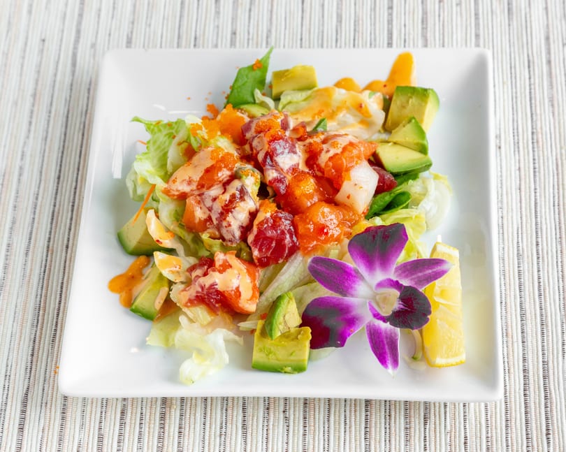 Colorful poke salad with avocado, lettuce, fish, and roe, garnished with a purple orchid and lemon.