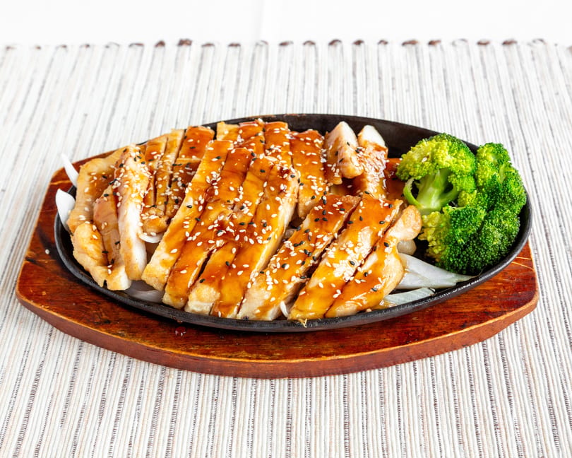 Teriyaki chicken with sesame seeds and broccoli on a sizzling plate with onions.