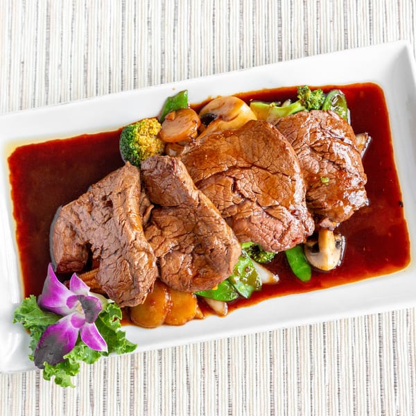 Four slices of beef in a rich brown sauce with broccoli, mushrooms, and snap peas, garnished with an orchid.