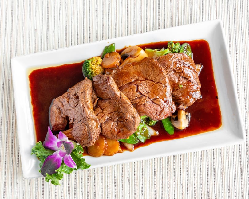 Four slices of beef in a rich brown sauce with broccoli, mushrooms, and snap peas, garnished with an orchid.