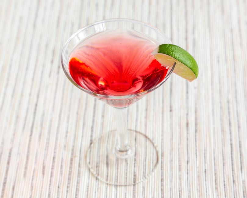 Vibrant red cocktail in a martini glass, garnished with a fresh lime wedge on a striped surface.
