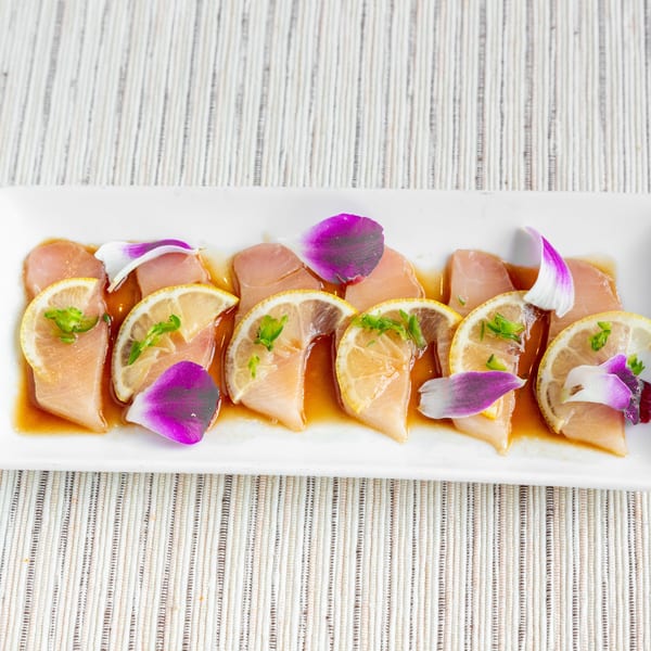 White rectangular plate with six pieces of fish sashimi, lemon slices, green herbs, and purple petals.