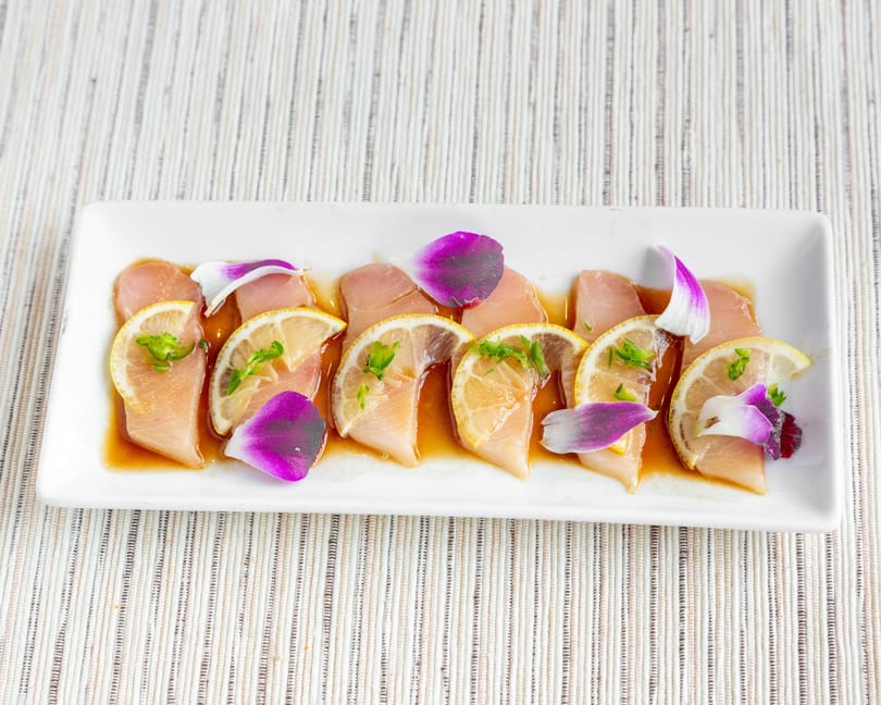 White rectangular plate with six pieces of fish sashimi, lemon slices, green herbs, and purple petals.
